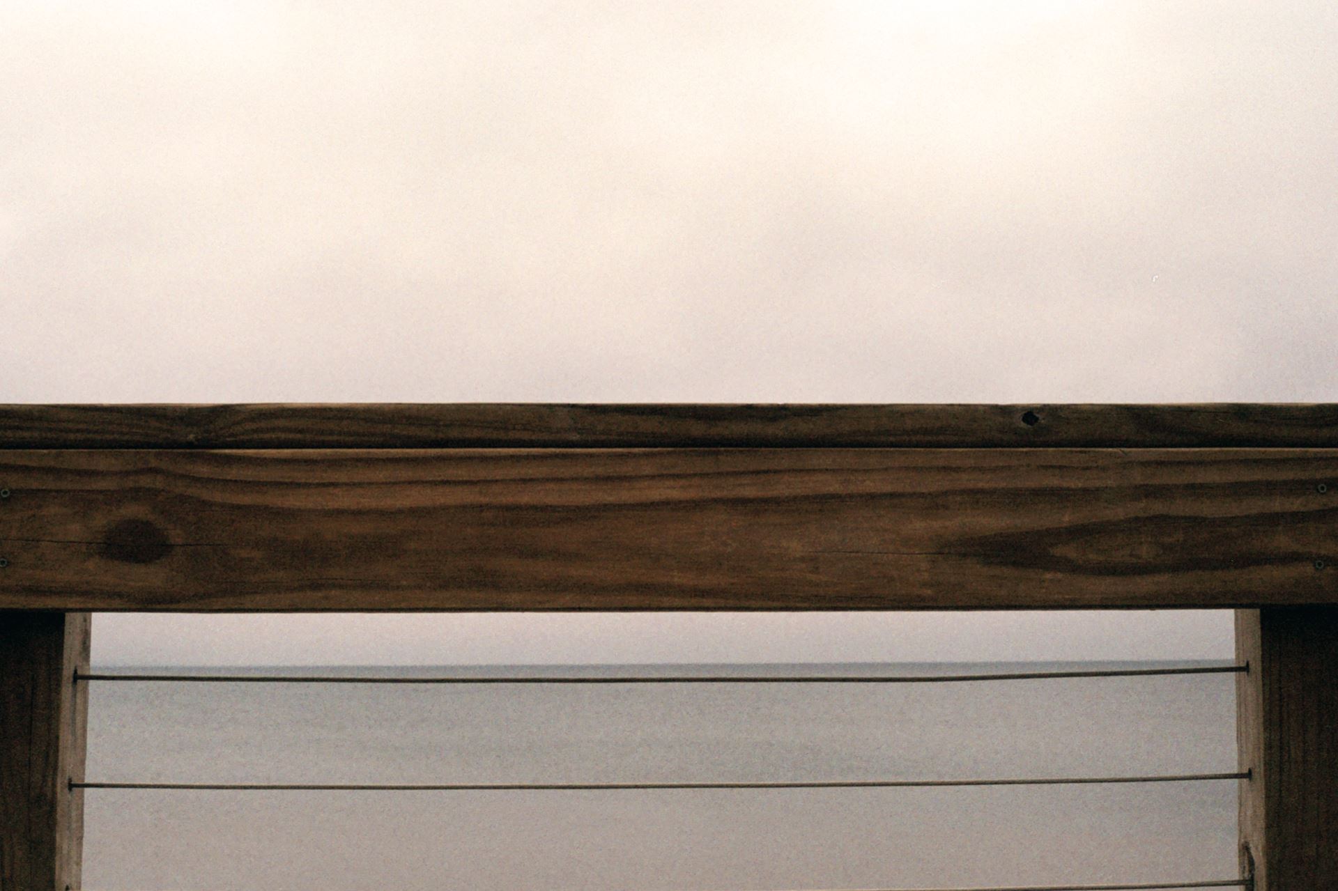 Close up section of wooden fence overlooking water and a gray sky. Emily Spaniol, “Beach Image 4,” film